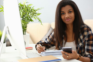 Smiling african american woman holding phone making notes in notebook checks, writes down important information