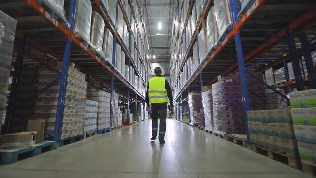 A worker walks through a factory warehouse. A man walks through the warehouse view from the back slowmotion. Rear view of a worker walking through a warehouse