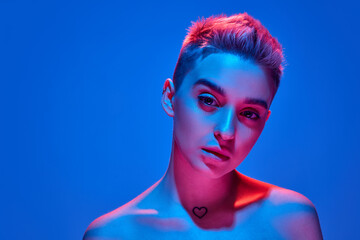Young beautiful girl with blonde short hair posing with bare shoulders against blue studio background with pink neon light. Deep attentive look. Concept of emotions, cyberpunk style, youth culture.