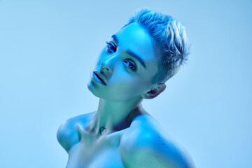 Beautiful young girl with short blonde hair and piercing posing with bare shoulders against blue...