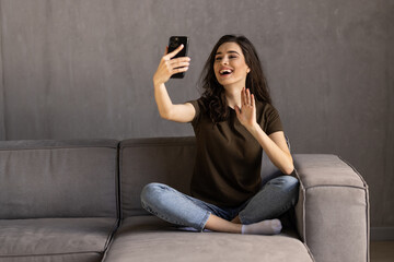 Smiling woman sitting on kitchen sofa talking by videocall dating online looking at phone. Video blogger vlogger recording vlog at home.