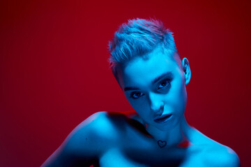 Uniqueness. Beautiful young girl with short blonde hair posing with bare shoulders against red studio background in blue neon light. Cyberpunk style. Concept of futurism, digital world, robot, art