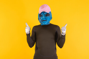 Man in blue headed alien face mask, wearing a pink cap, giving thumbs up. Concept of bizarre,...