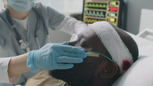 Selective focus close-up of doctor checking and fixing bandage and nasal cannula for unconscious African American man with head injury