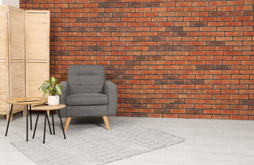 Cosy armchair and potted plant on coffee table near brick wall in room, space for text. Interior design