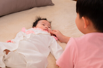 Selective focus of Asian newborn baby lying down on bed play with elder brother, boy playing with sister hold baby cheeks affectionately and teasing with tenderness. happy family concept.