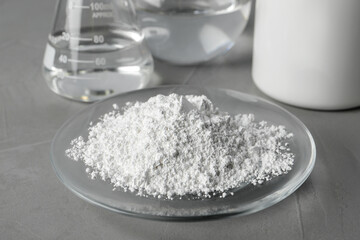 Plate with calcium carbonate powder and laboratory glassware on grey table, closeup