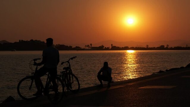 Silhouette of a man with bicycles at sunset. Silhouettes of two men with bicycles on the sea promenade taking pictures on the phone at a summer sunset.