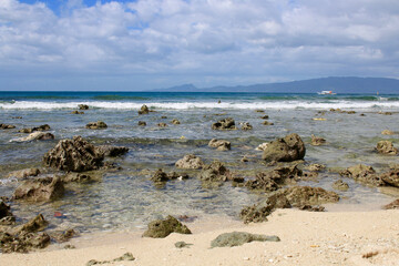Fototapeta na wymiar Sandy beach of a tropical island. Overcast weather. Sea wave rolls on the sandy beach and stones near the shore.