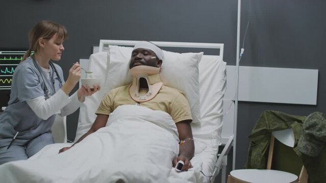 Professional nurse working in modern military hospital spoon feeding injured Black serviceman