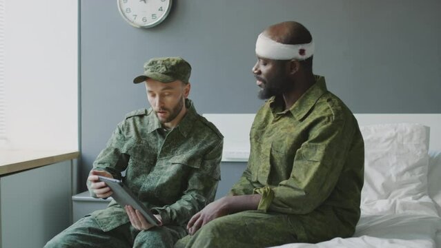 Caucasian lieutenant visiting his injured Black colleague in military hospital showing him news on digital tablet