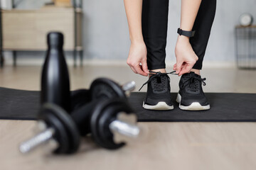Fototapeta na wymiar sport and healthy lifestyle concept - woman preparing for training at home and tying shoelaces on sneakers - dumbbells and bottle of water on first plan