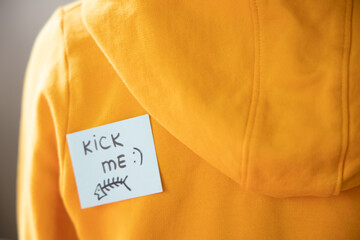 Boy in yellow hoodie with a sticky note on his back, saying kick me.