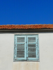 blue wooden shutters with peeling  paint against white cement rendered wall with old roof tiles and clear blue sky