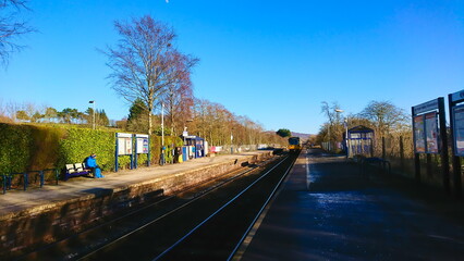 Hope Station, Peak District - February 25, 2018: passengers waiting for a train on the platform in a typical rural British railway station while a train approaches under a clear blue sky