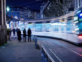 A city night scene with a group of people illuminated by the passing train, creating blurred motion in an urban area filled with built structures and various modes of transportation.
