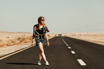The girl is standing on the highway in the middle of the desert. The model is dressed in shorts, a shirt and sneakers. The woman is smiling and her hair is blowing in the wind. Black asphalt