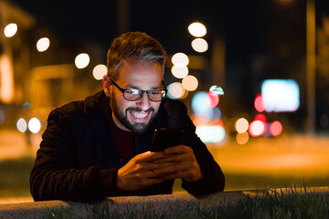 A happy man with glasses is using a smartphone in the city with a nightlight in the background.