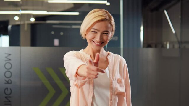 Playful cheerful blonde office worker wearing a white t-shirt playfully beckons the viewer to join in on the fun by pointing her finger at the camera and saying Hey, come over here, while dancing.
