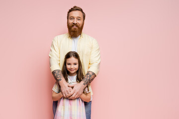 Cheerful man hugging preteen child and looking at camera isolated on pink.