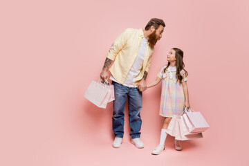 Tattooed man holding hand of daughter with purchases on pink background.