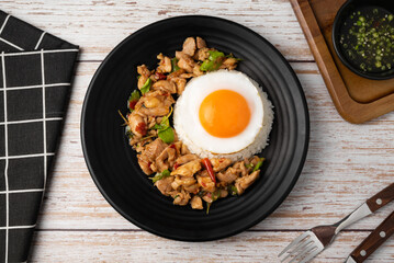 Top view of stir fried basil and chicken on rice with fried egg in black dish on wooden table.
