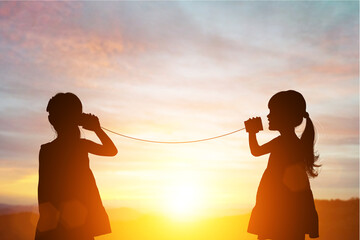 Silhouette of Two female kids stands and talking by using string can phone on beautiful sunset sky - obrazy, fototapety, plakaty