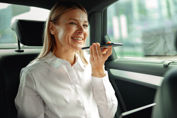 Businesswoman seated in car backseats recording voice message on phone on the way to work