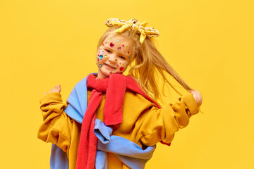 Portrait of little girl wearing bow on head touching blond hair and smiling to camera over yellow background