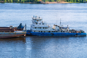 A tugboat pushes a large barge with cargo along a wide river. In summer, the tugboat goes down the river. Cargo transportation by river. Navigation of vessels on the river.