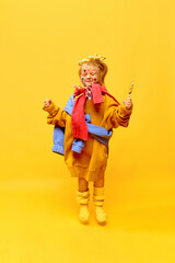 Full-length photo of little girl, jumping with lollipop and smiling with closed eyes over yellow background. Concept of fashion, ad, children's products, childhood, child model