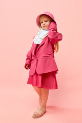 Photo of little, stylish girl touching headdress and wearing big pink, oversized clothes to imitate her mother over pink background. Full-length