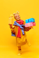 Full-length photo of little girl holding gifts, boxes with happy face over yellow background. Concept of celebration, ad, children's products, childhood, family