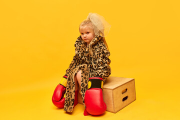 Little upset blond girl wearing boxing gloves and leopard coat sitting on boxes and looking away...