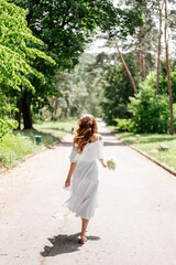 A slender beautiful girl in a white dress with a white bouquet in her hands walks cheerfully along the park path. Her dress and hair fly in the wind