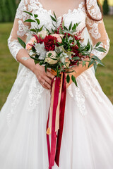 A bouquet of burgundy chrysanthemums and delicate pink roses, decorated with green leaves and red ribbons, in the hands of a bride in a white dress with lace, on a blurred background