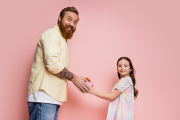 Cheerful bearded father and preteen girl holding gift box and looking at camera on pink background.