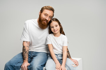 Positive tattooed man sitting near daughter on cube isolated on grey.