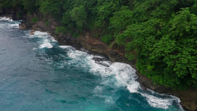 Static aerial view of tidal waves splashing on brown rocks with green trees. Amazing copter footage of turquoise sea water washing rocky island. Idyllic tropical scene. Stormy ocean at wild lagoon.