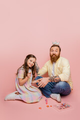 Positive girl holding nail polish near tattooed dad with crown on head and decorative cosmetics on pink background.