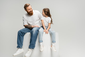 Positive tattooed man looking at daughter in t-shirt and jeans sitting on cube isolated on grey.