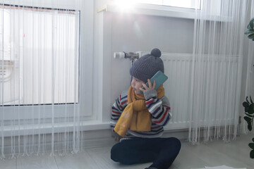The child is warmly dressed in a sweater and a hat, sitting near the heater with bills in her hands, money and a phone. The concept of a crisis and a large set for utilities.