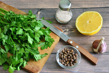 A cutting board with a knife and bunch of parsley, allspice, salt shaker and half of lemon