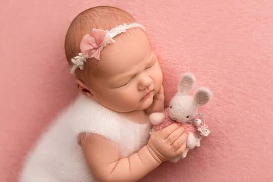 Top view of a newborn baby girl sleeping in a white overalls, with a white bandage on her head. With a knitted white rabbit on a pink background. Beautiful portrait of a newborn baby 7 days, one week.