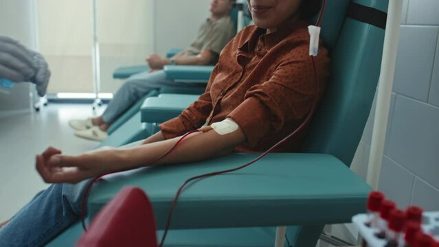 Medium section shot of young adult Hispanic woman sitting in armchair in modern hospital donating her blood, medical worker checking the process