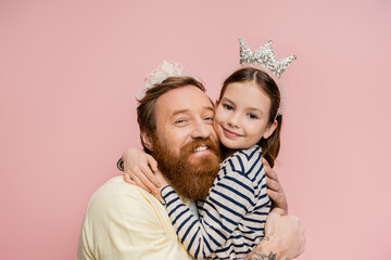 Positive father and preteen girl in crown headbands hugging isolated on pink.