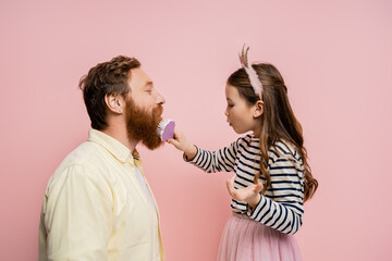Side view of preteen girl with crown headband brushing beard of father isolated on pink.