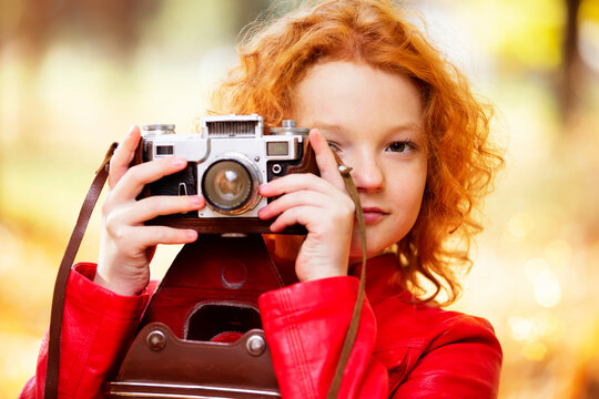 Little red-haired girl with a retro camera on an autumn background.