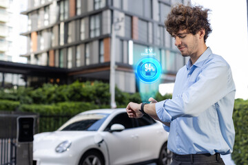 Progressive businessman check wearable hologram watch on electric car battery that being recharge at charging station point in the city. Digital interface, data from EV car display on smartwatch.
