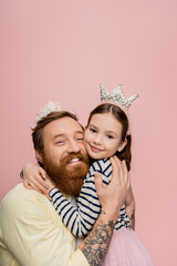 Smiling tattooed man in crown headband hugging daughter isolated on pink.
